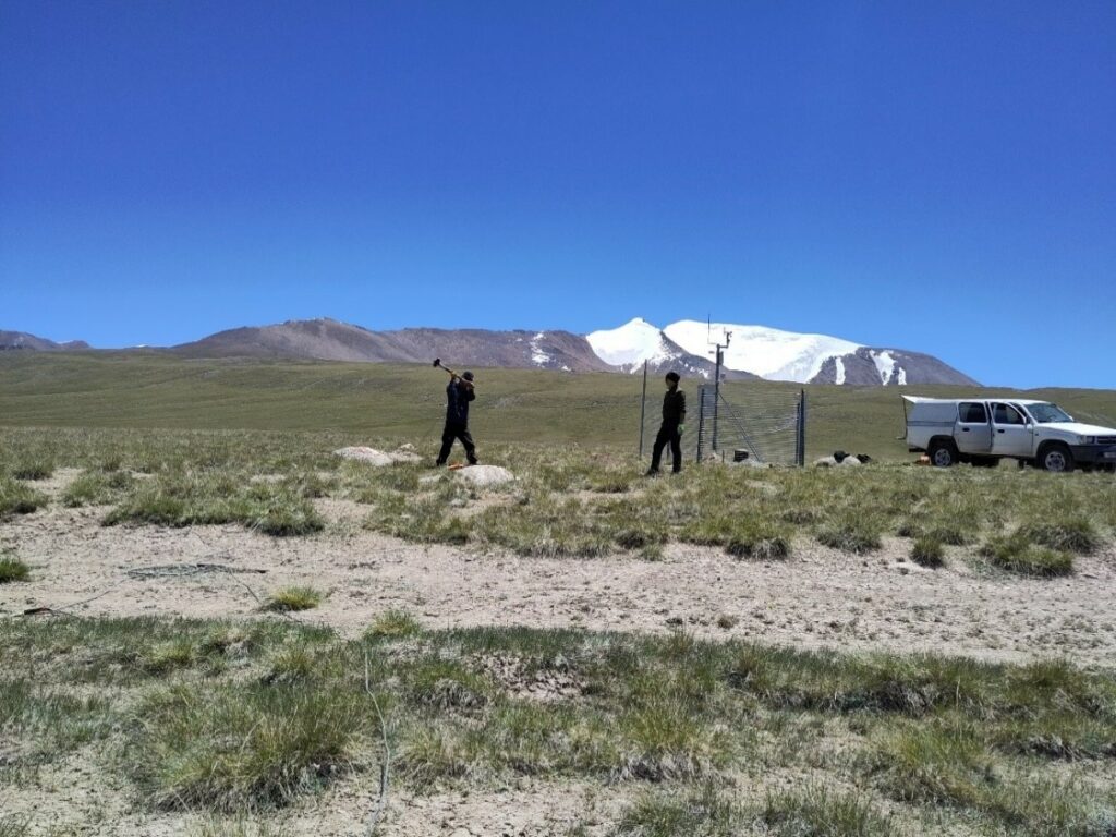 A grassy landscape with mountains in the background. Two people are installing a scientific instrument with fences. There is a white pick-up truck on the right.
